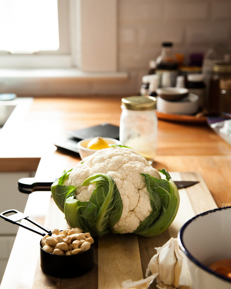 Image shows a head of cauliflower on top of a cutting board in a kitchen scene.