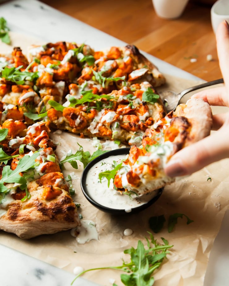 Image shows a hand dipping a slice of vegan Buffalo cauliflower pizza into a small bowl of creamy sauce.