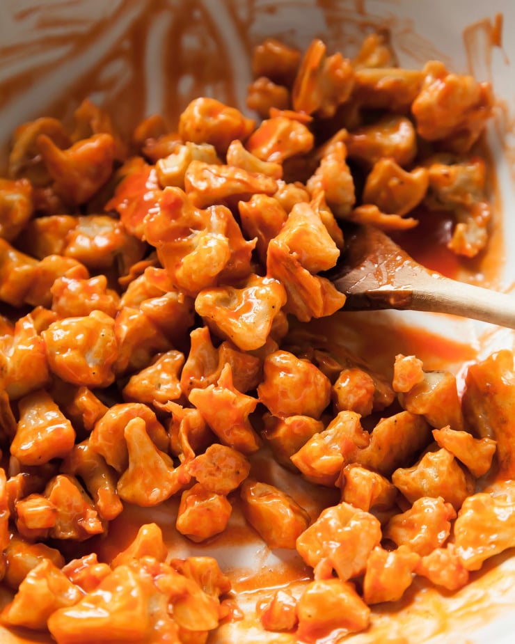 Image shows battered cauliflower being tossed in a bright orange Buffalo sauce.