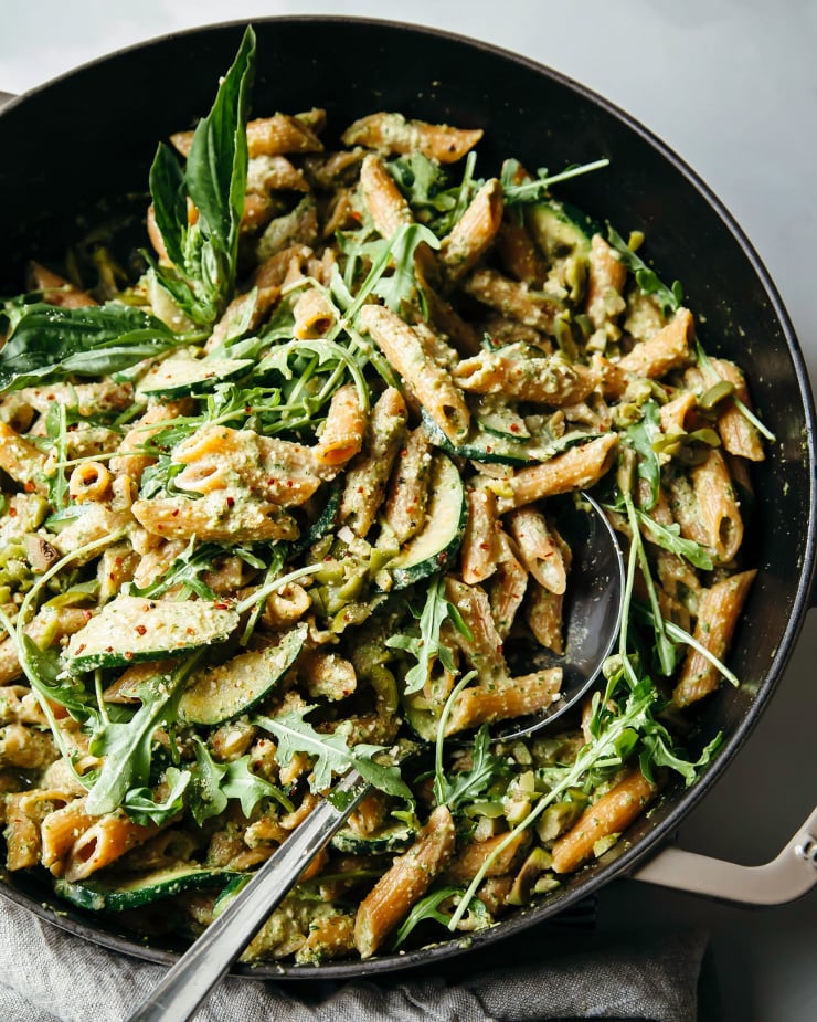 An overhead shot of a penne pasta dish with arugula, cooked slices of zucchini, ground chilies, and olive pesto; all in a braiser-style pot with a ladle sticking out for serving.