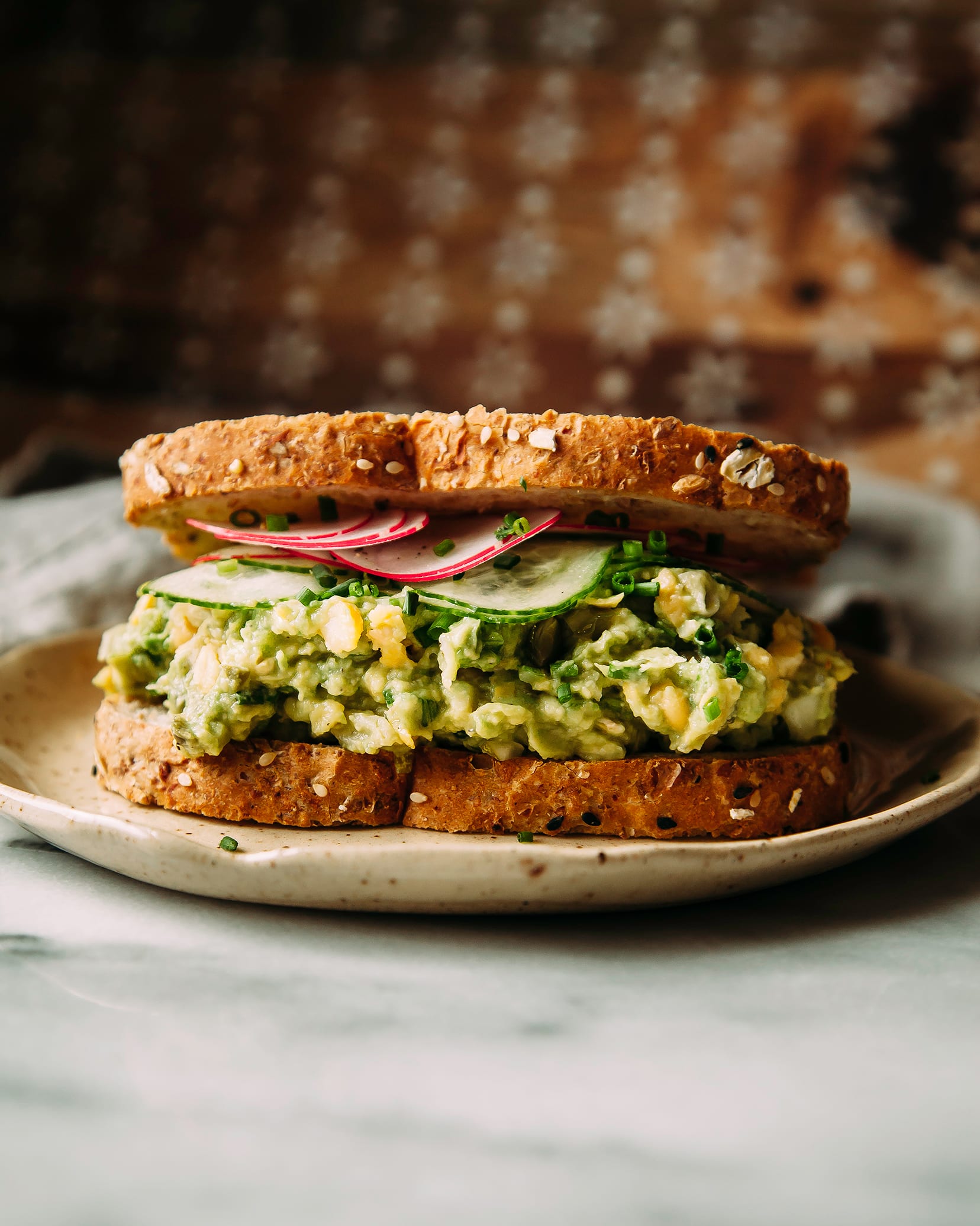 A head-on shot of a sandwich filled with avocado chickpea salad, sliced radishes, and cucumbers. The bread is grainy. The sandwich is on a beige plate on a grey marble surface.