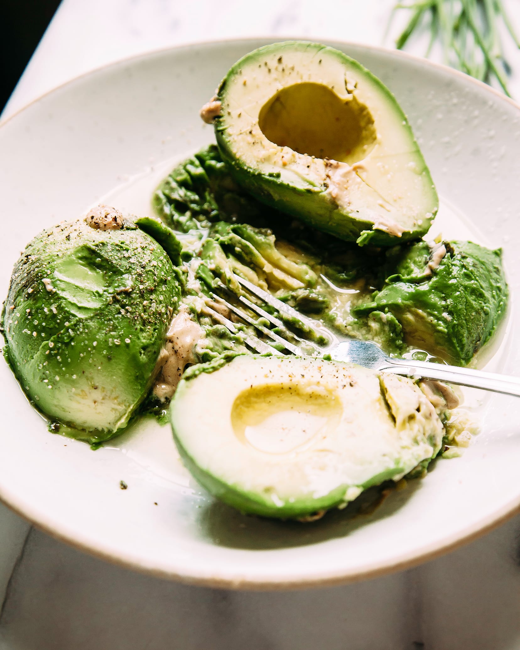 A 3/4 angle shot of avocado halves i a white bowl getting mashed with seasonings.