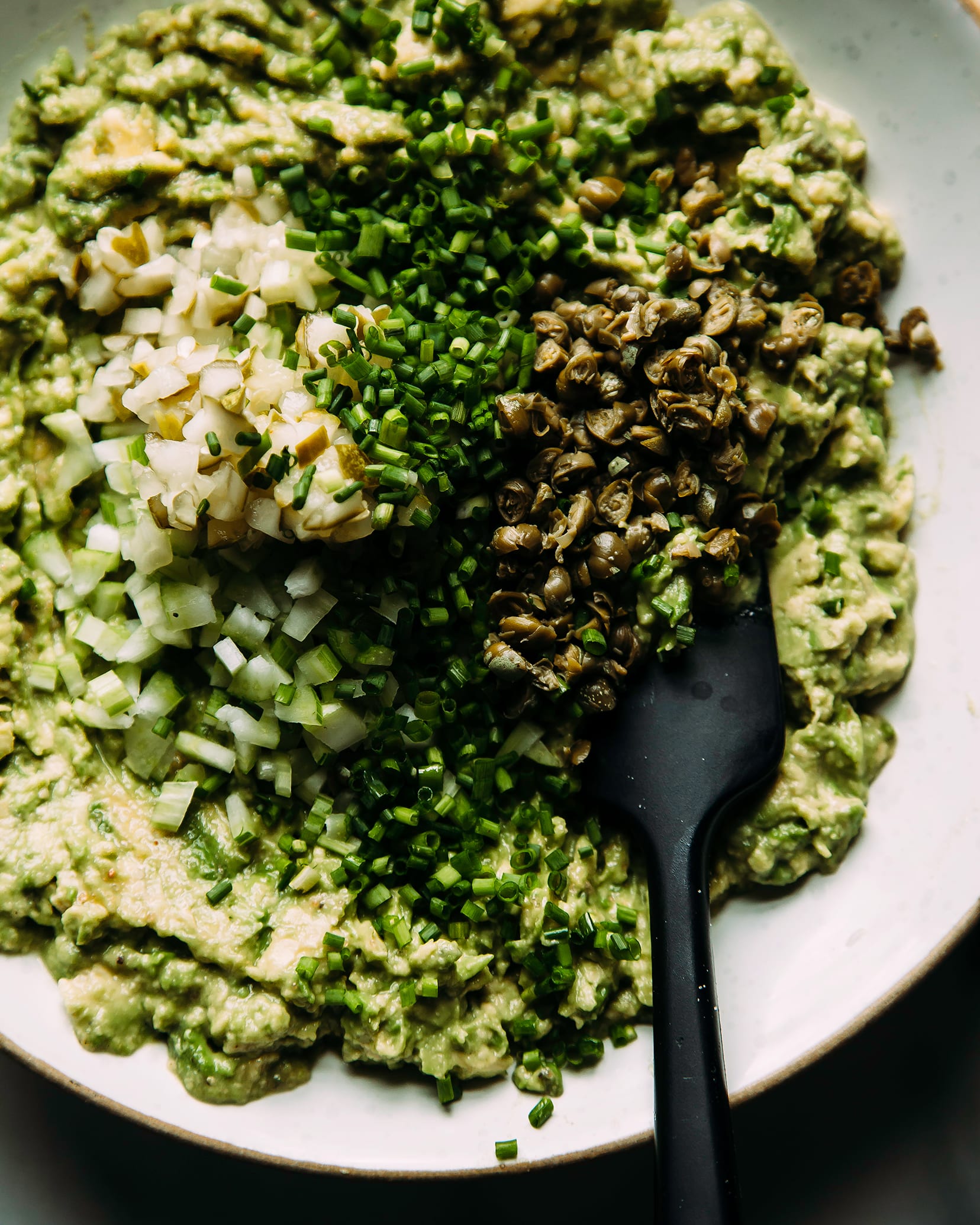 An overhead shot of avocado chickpea salad in a white bowl with capers, chives, pickles and celery (all very finely minced)