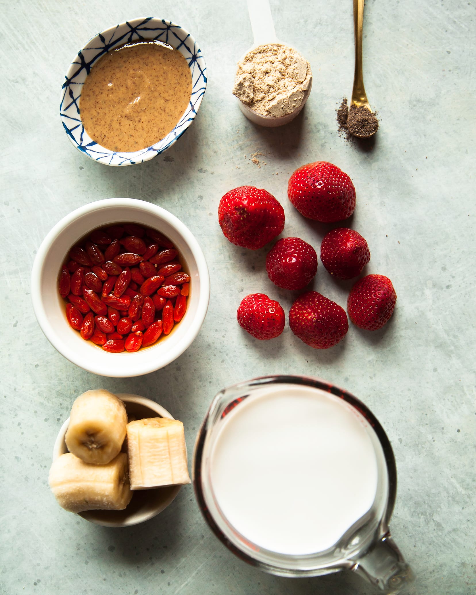 An overhead shot of ingredients for a strawberry goji berry smoothie on a light blue, painted background.
