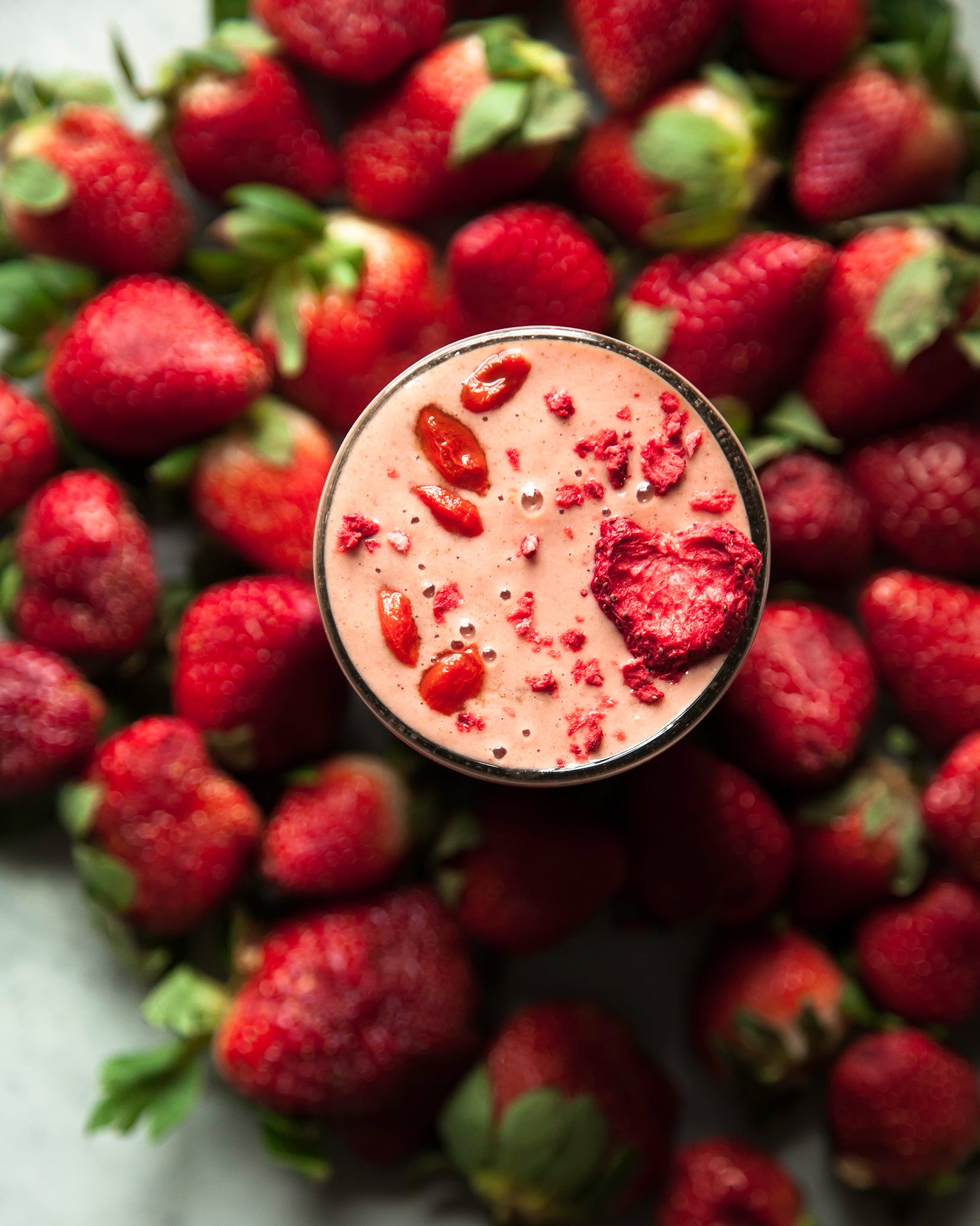 Overhead shot of a light pink strawberry goji smoothie, surrounded by fresh strawberries. It is garnished with soaked goji berries and a slice of dehydrated strawberry.