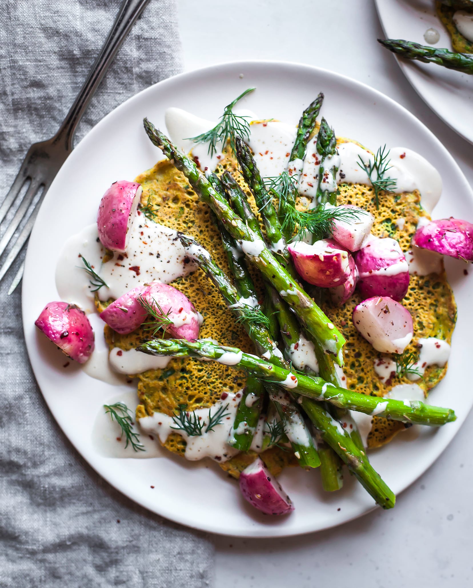 Image is an overhead shot of a yellow chickpea flour pancakes topped with a creamy white sauce and roasted vegetables. The dish is on a white plate with a grey linen napkin nearby.
