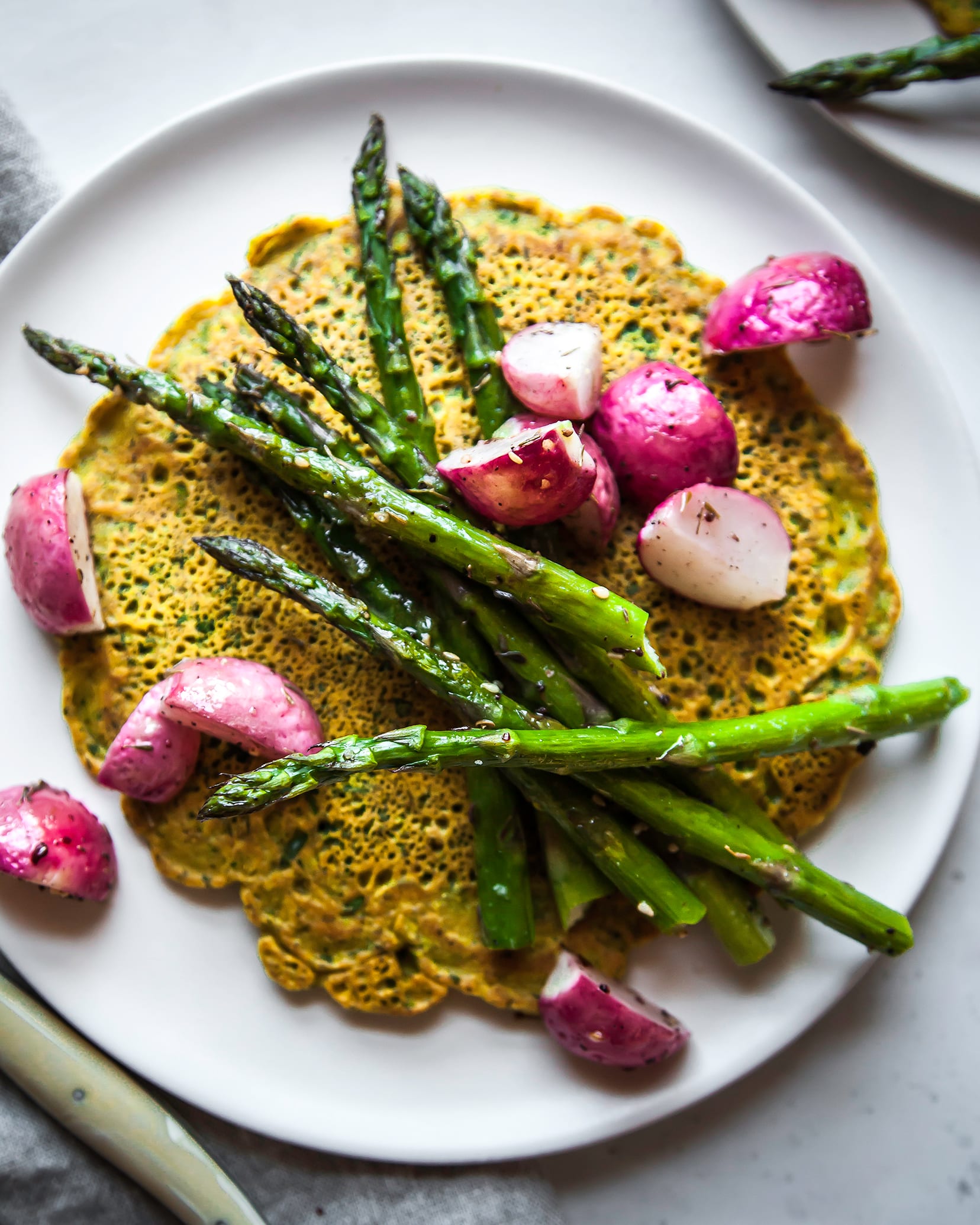 Image is an overhead shot of a yellow chickpea flour pancakes topped with roasted vegetables. The dish is on a white plate with a grey linen napkin nearby.