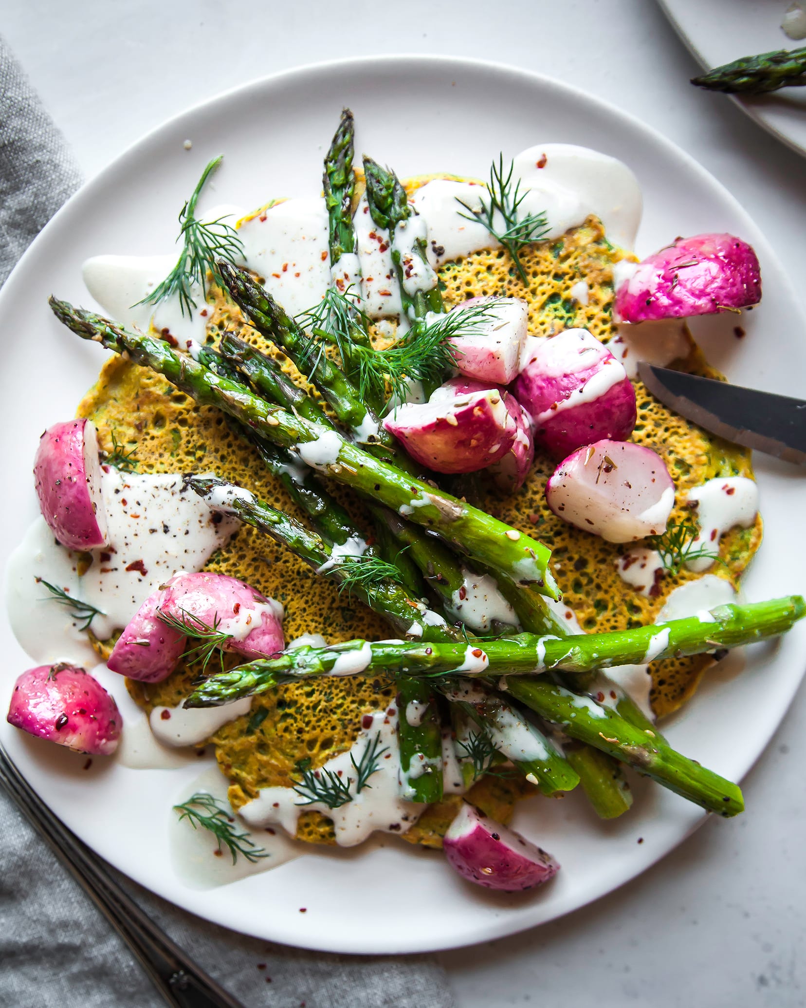 Image is an overhead shot of yellow chickpea pancakes topped with a creamy white sauce and roasted vegetables. The dish is on a white plate with a grey linen napkin nearby.