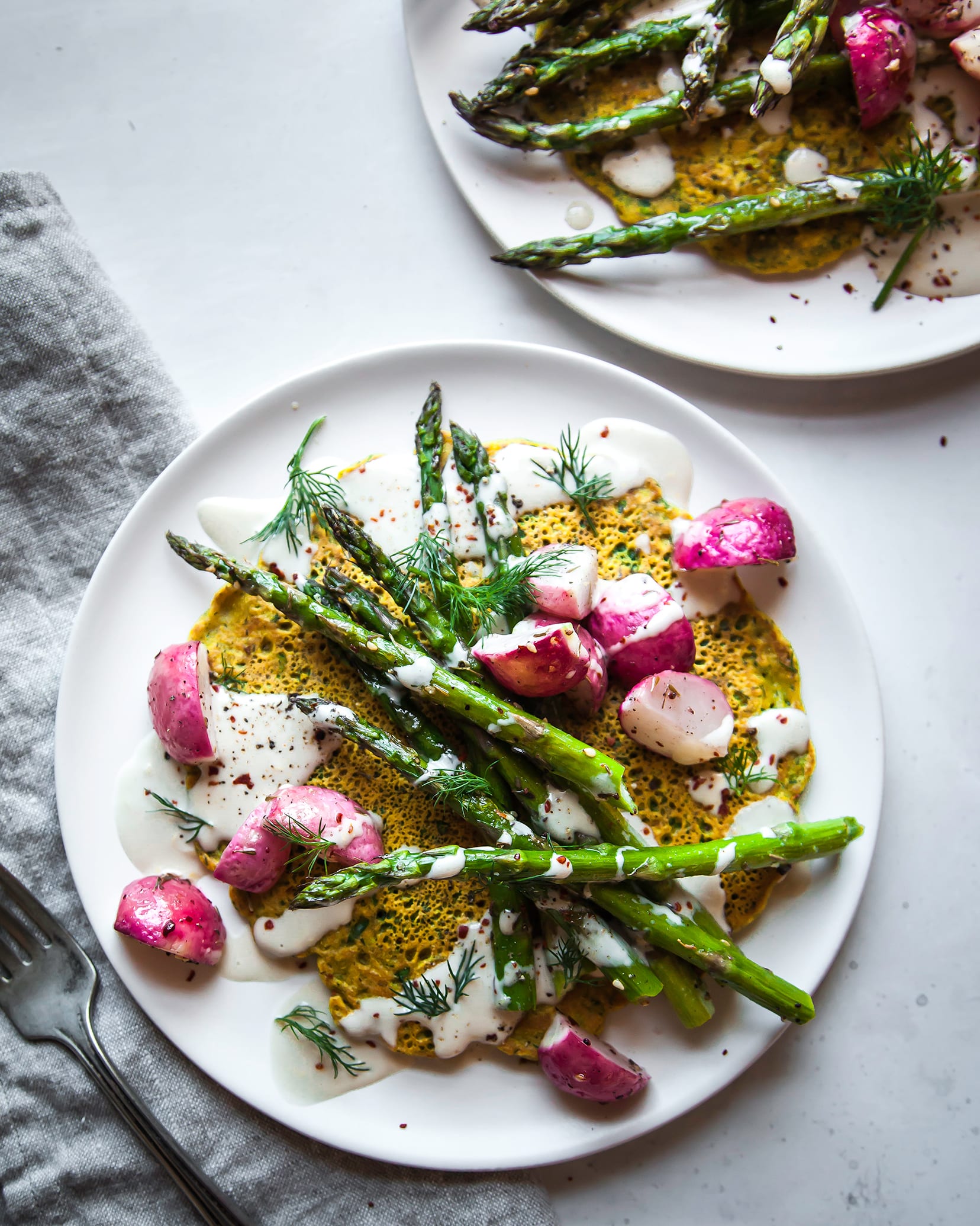 Image is an overhead shot of a yellow chickpea flour pancake topped with a creamy white sauce and roasted vegetables. The dish is on a white plate with a grey linen napkin nearby.