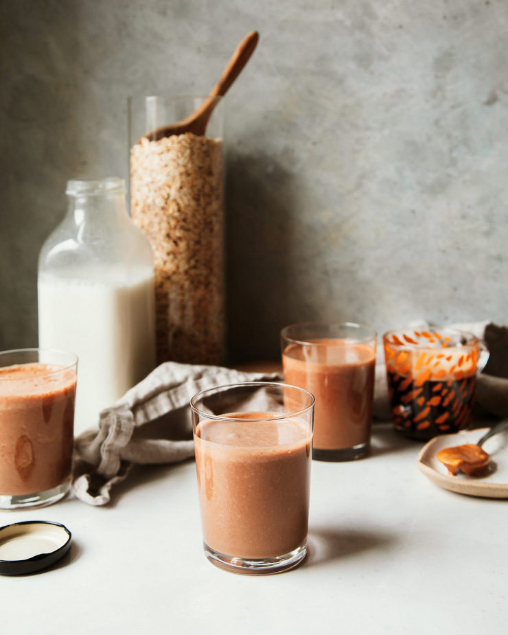 A head on shot of a thick peanut butter mocha smoothie that is rich, chocolate brown in colour. In the background, there is a clear bottle of homemade non-dairy milk and a big clear jar of oats.