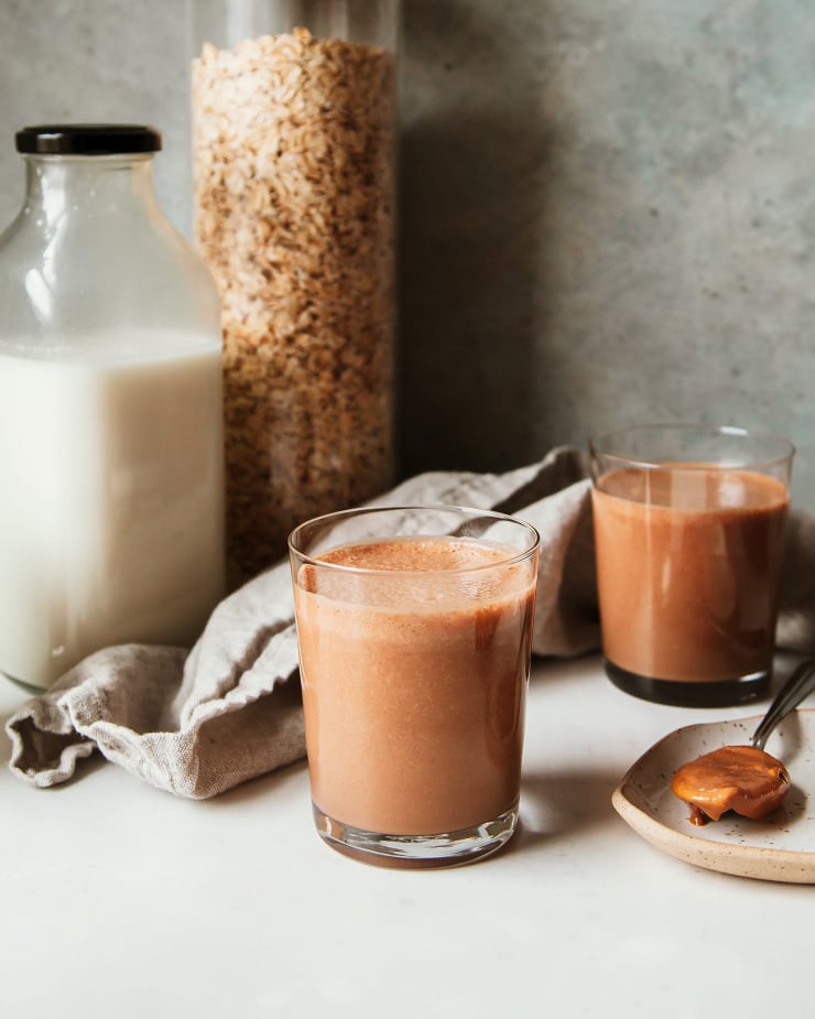 A head on shot of a thick peanut butter mocha smoothie that is rich, chocolate brown in colour. In the background, there is a clear bottle of homemade non-dairy milk and a big clear jar of oats.