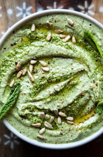 An up close, overhead shot of a creamy, light green dip. The dip is garnished with sunflower seeds and olive oil.
