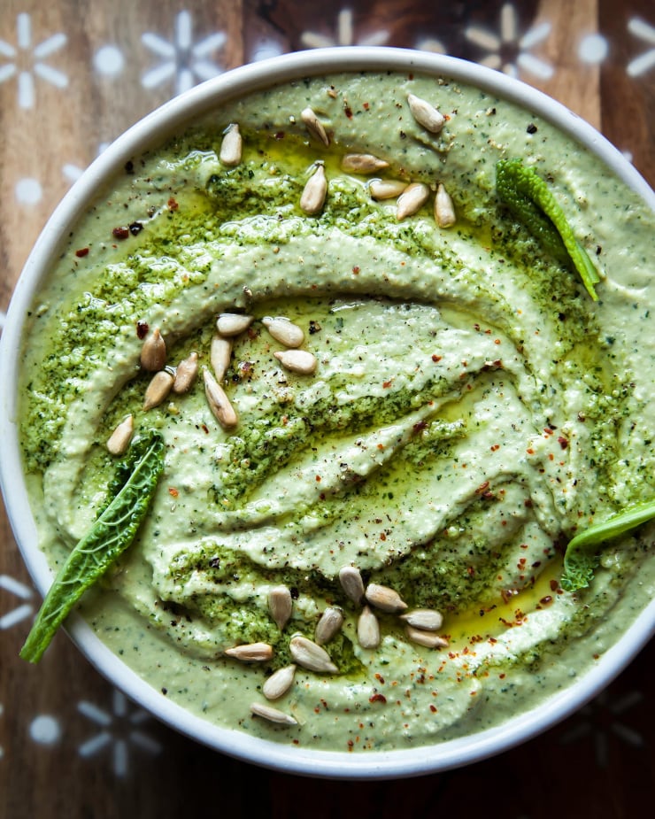 An up close, overhead shot of a creamy, light green dip. The dip is garnished with sunflower seeds and olive oil.