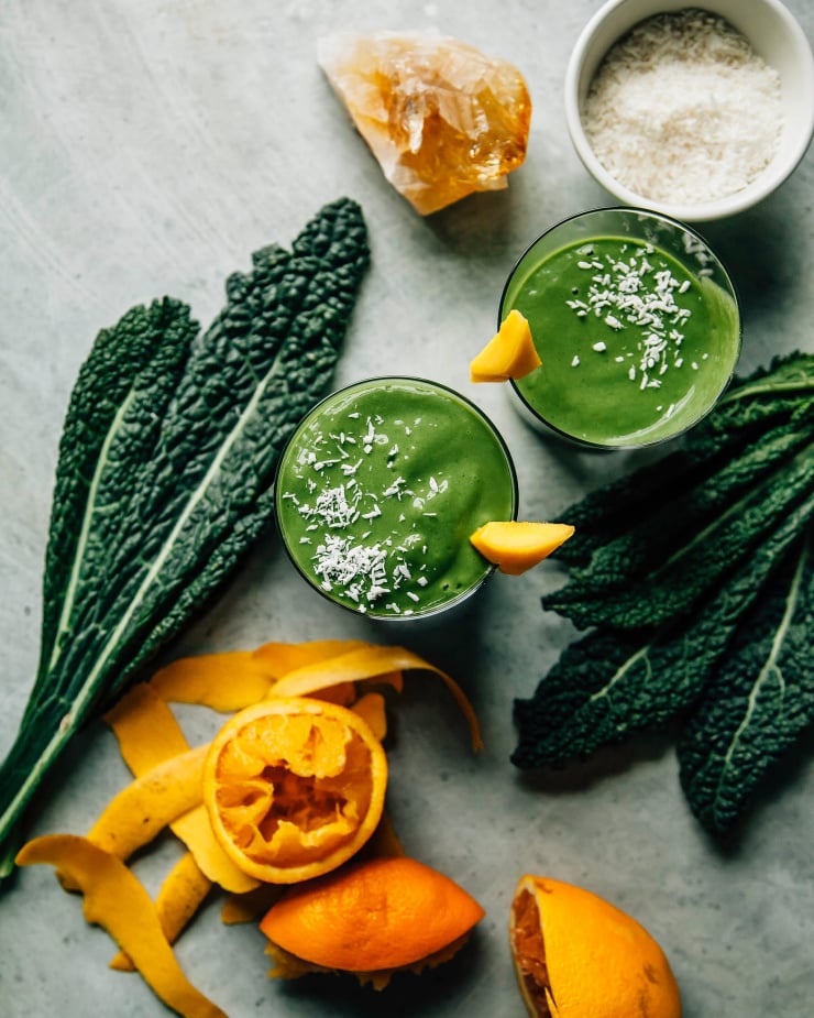 Overhead shot of 2 deep green smoothies in clear glasses on a light blue background. Kale leaves and squeezed oranges are nearby. The smoothies are garnished with shredded coconut and a piece of mango is perched on the rim of the glasses.