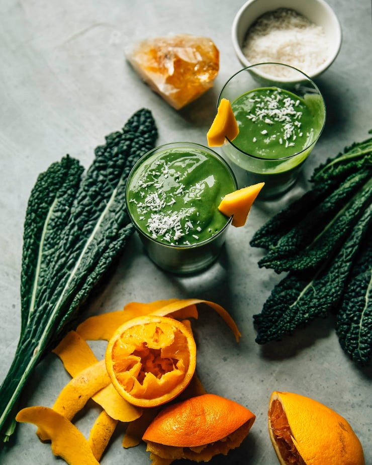 Overhead shot of 2 deep green smoothies in clear glasses on a light blue background. Kale leaves and squeezed oranges are nearby. The smoothies are garnished with shredded coconut and a piece of mango is perched on the rim of the glasses.