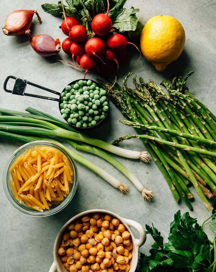 An overhead shot of ingredients for a pasta salad with Spring vegetables over a speckled light blue background.