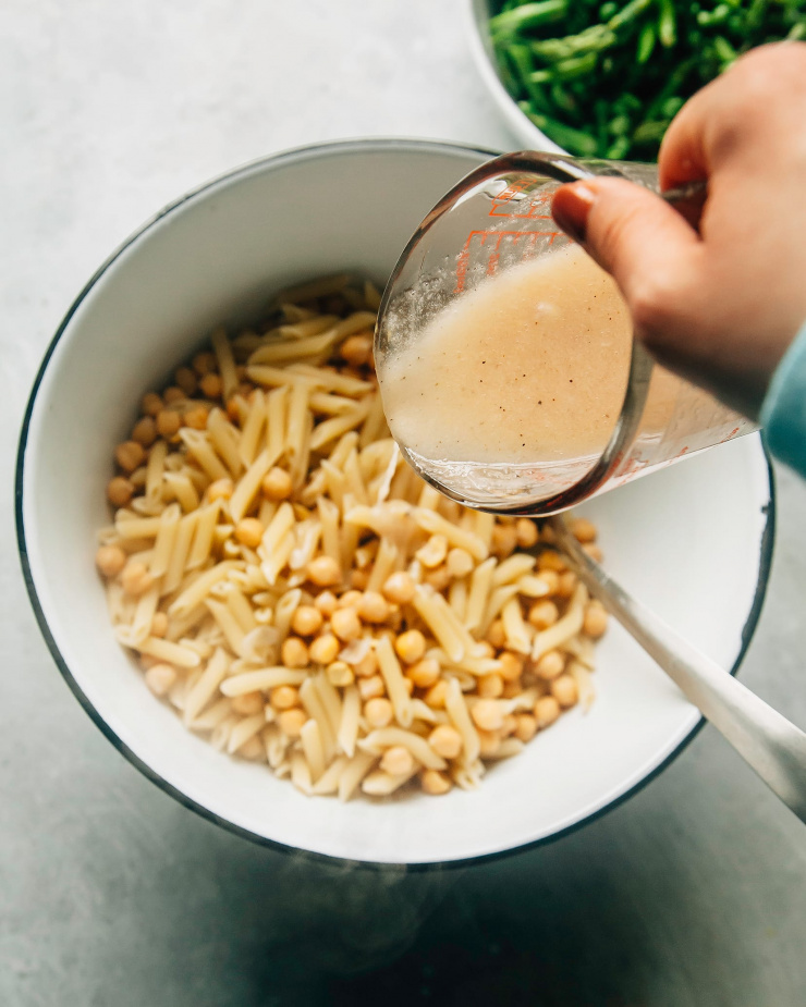A hand is pouring vinaigrette over some cooked pasta and chickpeas in a white bowl with a black rim.