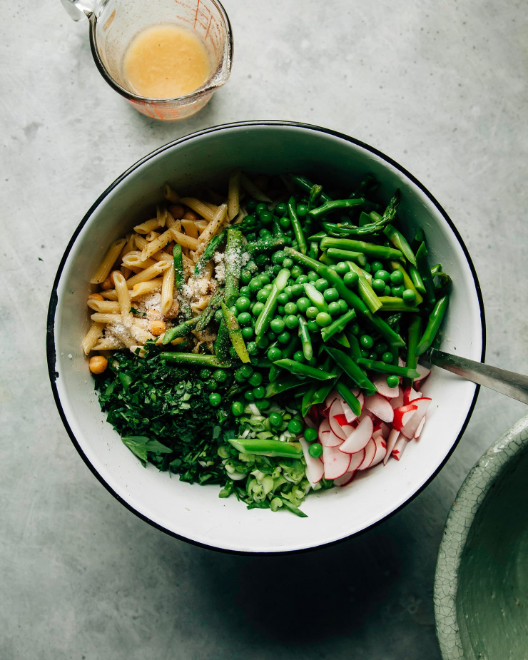 An overhead shot of a bunch of green vegetables, cooked pasta and chickpeas in a white bowl with a black rim. There is a glass measuring cup of vinaigrette nearby.