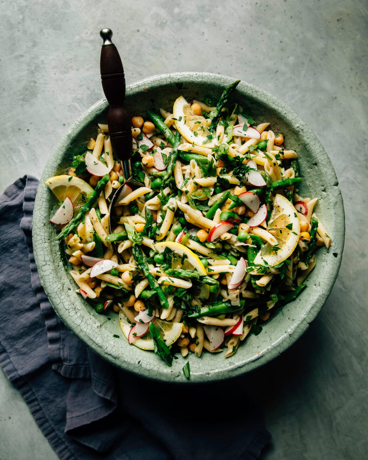 An overhead shot of a pasta salad with lots of green vegetables and herbs in a sage-green ceramic bowl.
