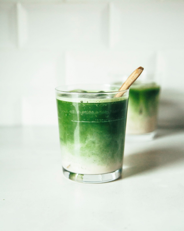 Photo shows a head on shot of a creamy, green beverage in a clear glass against a white background.