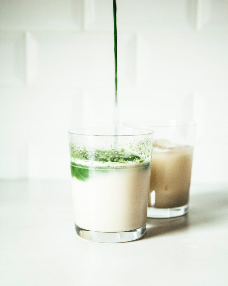 Photo shows a dark green liquid being poured into a creamy glass of oat milk.