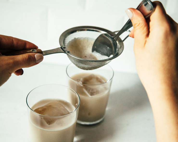 Photo shows two hands straining some oat milk into a glass with some ice in it.