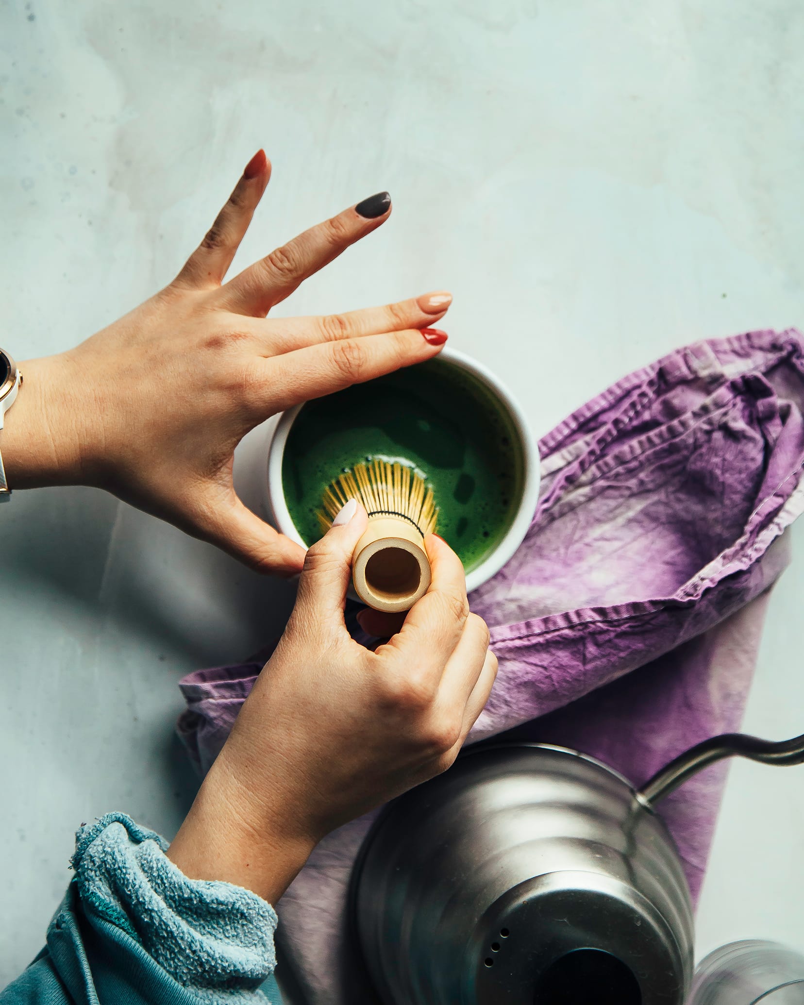 Photo shows a pair of hands whisking some matcha in a small white bowl with a bamboo whisk. There is a purple napkin nearby.