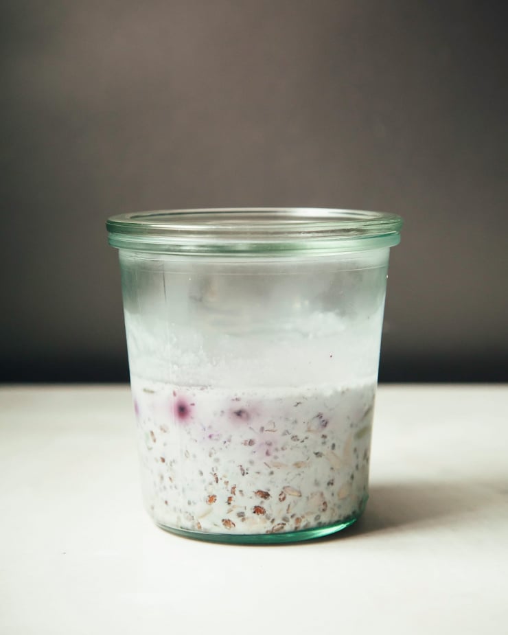 A head-on photograph of a jar with mixed seeds, oats, and frozen blueberries.