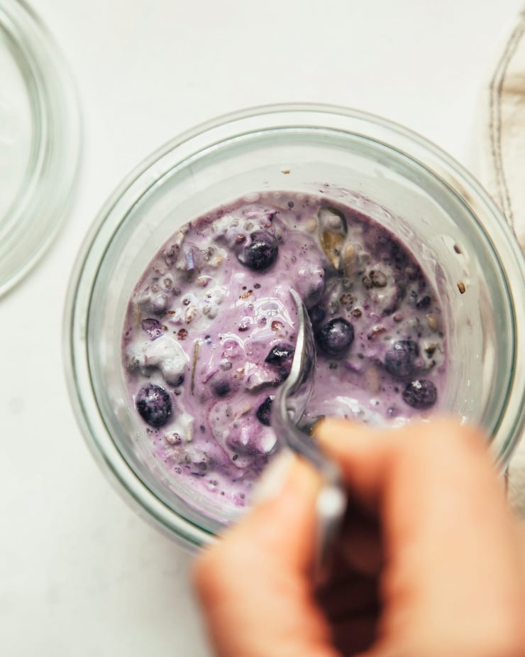 A hand stirs together frozen blueberries, oats, chia, seeds and coconut in a glass jar.