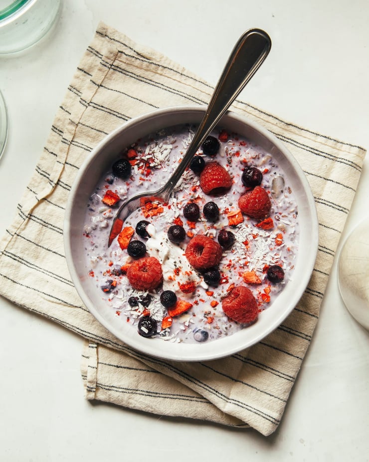 An overhead shot of light purple Bircher muesli in a white bowl over a striped napkin. The muesli is topped with fresh berries and shredded coconut.