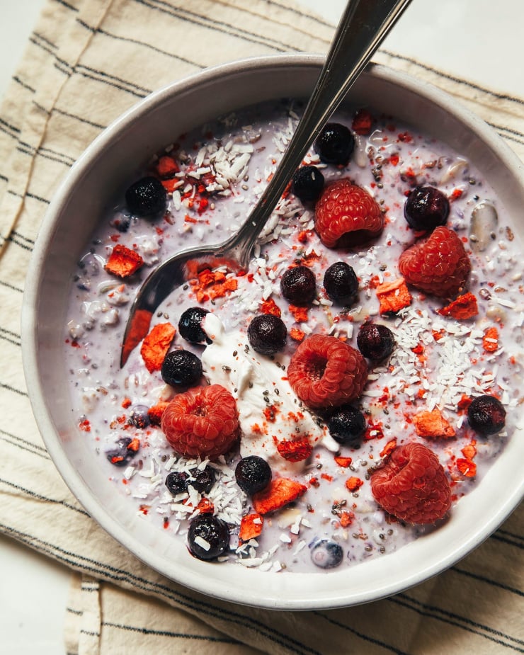 An overhead shot of light purple Bircher muesli in a white bowl over a striped napkin. The muesli is topped with fresh berries and shredded coconut.