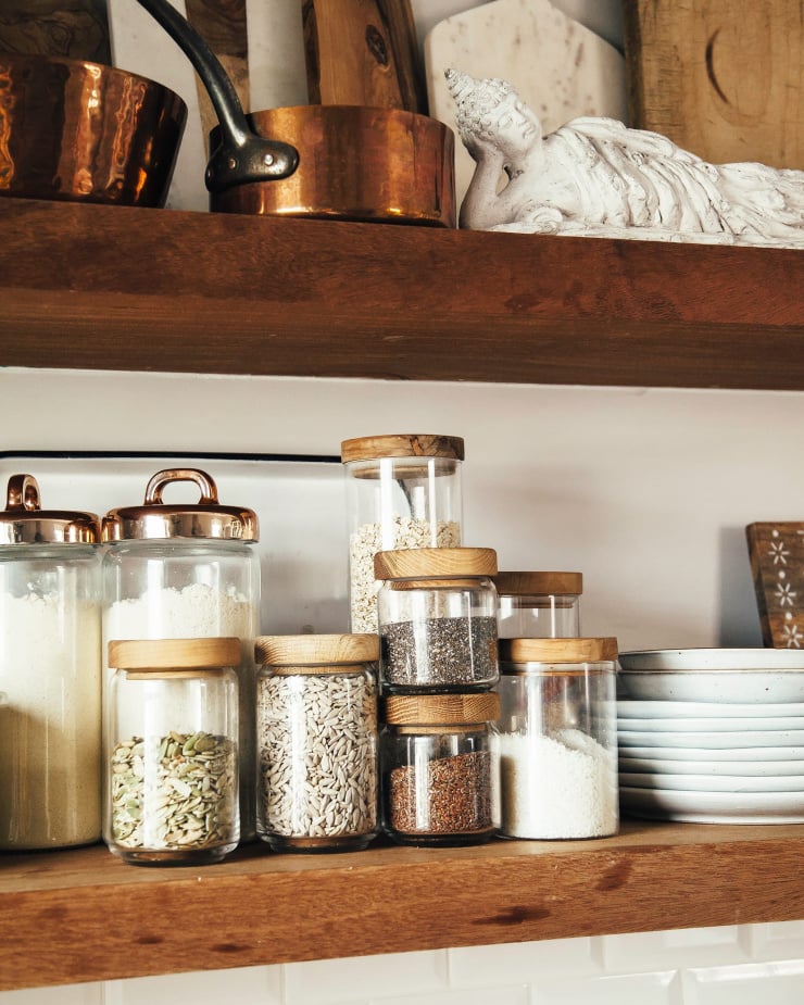 The image shows open shelving in a kitchen with a focus on storage jars filled with pantry staples like oats, seeds, and shredded coconut.