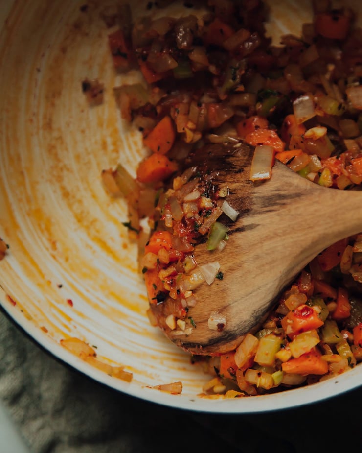 A wooden spoon is stirring sautéed vegetables at the bottom of a pot.