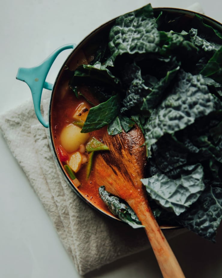 Image shows chopped kale being added to a pot of stew.