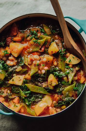 An overhead shot of smoky and spicy white bean stew in a turquoise pot on a beige linen background. The red-hued stew is chunky and loaded iup with white beans, wilted kale, cross cuts of Romano beans, and is topped with chili flakes.
