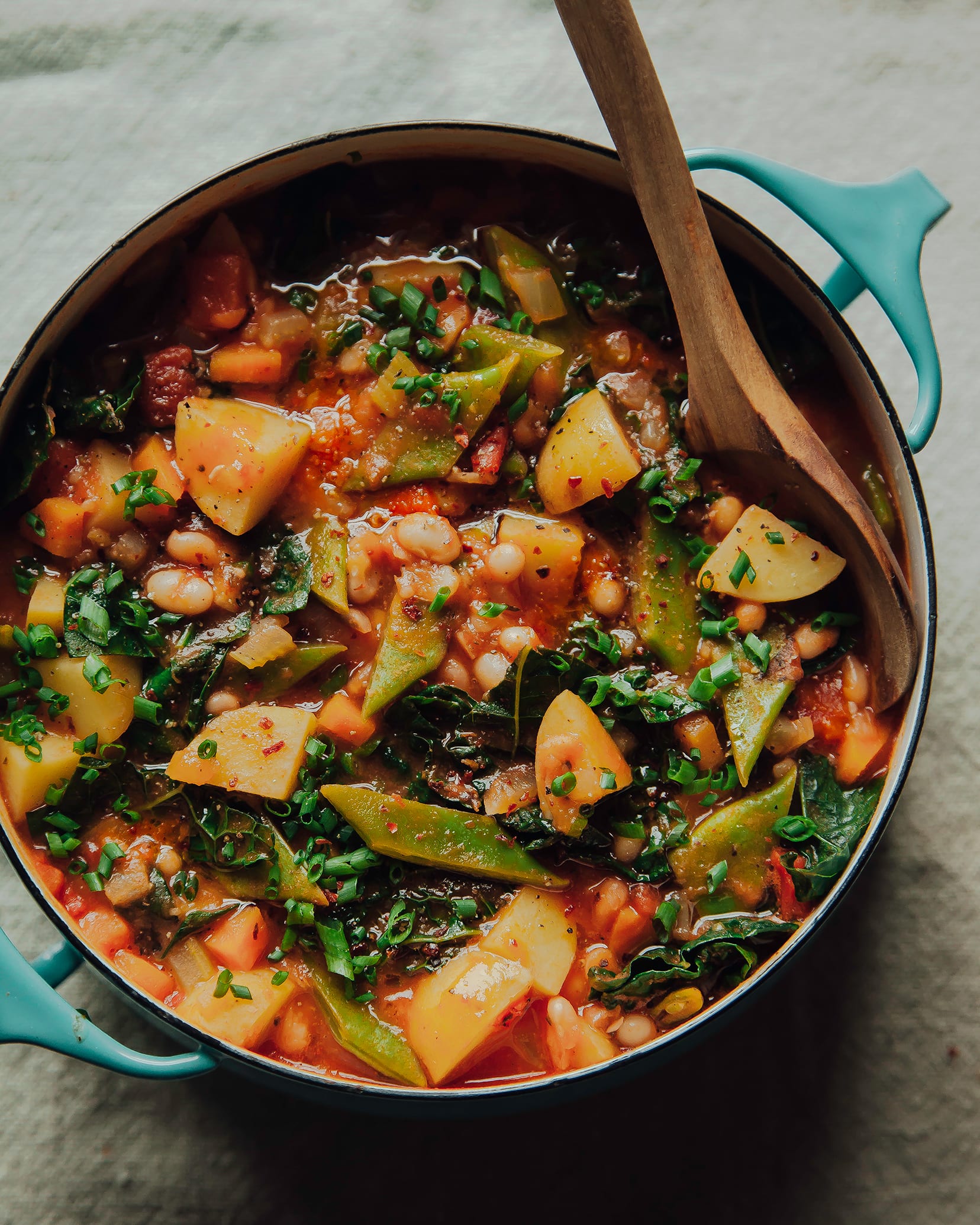 An overhead shot of smoky and spicy white bean stew in a turquoise pot on a beige linen background. The red-hued stew is chunky and loaded iup with white beans, wilted kale, cross cuts of Romano beans, and is topped with chili flakes.