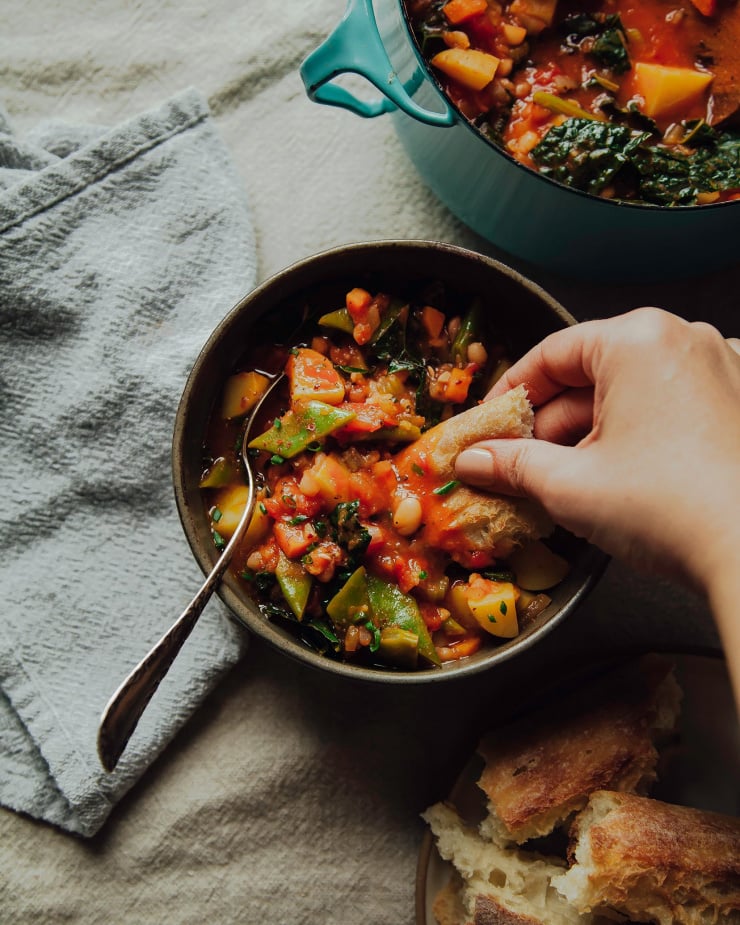Image shows a hand dipping a piece of bread into a bowl of a deep red white bean soup.