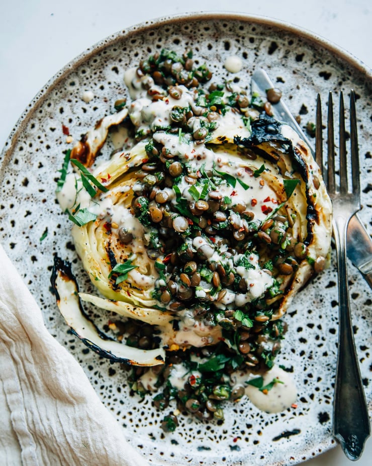 Image shows a grilled cabbage steak topped with lentils, herbs and a creamy sauce.