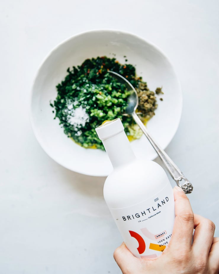 A hand is pouring olive oil into an herb-based sauce.