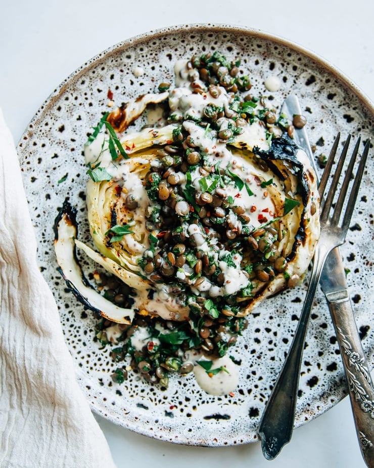 Image shows a grilled cabbage steak topped with lentils, herbs and a creamy sauce.