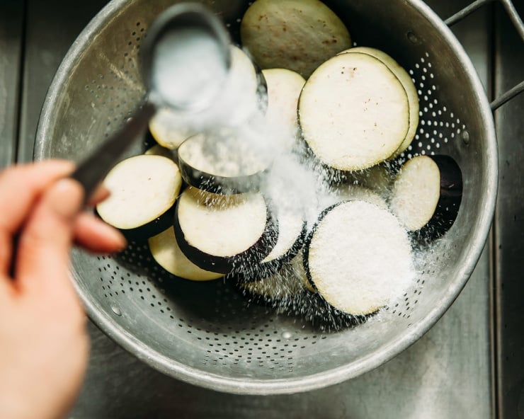 An overhead shot of eggplant slices in a colander. A hand is spooning salt all over the eggplant.