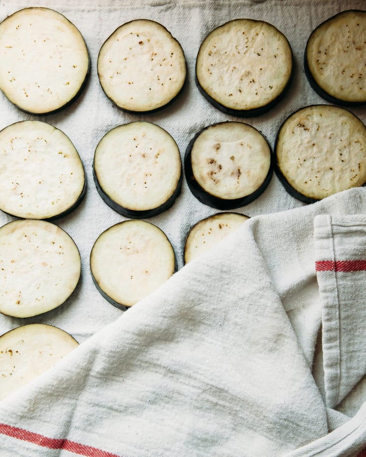 An overhead shot of eggplant slices being blotted with a dish towel.