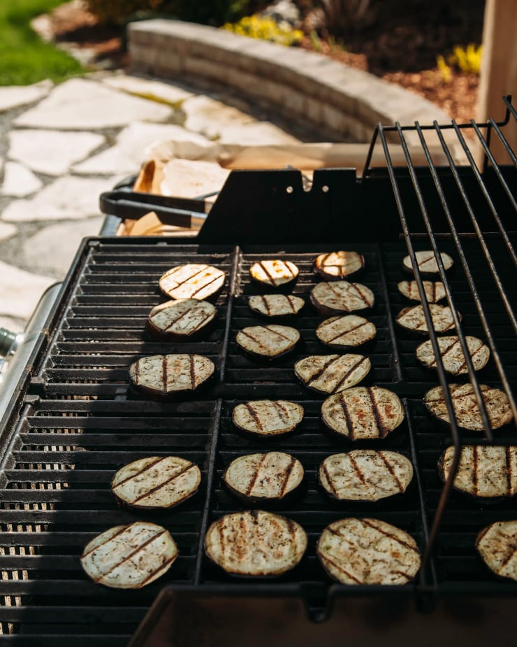 A 3/4 angle shot of a grill outdoors with a bunch of eggplant slices being cooked on the grates.