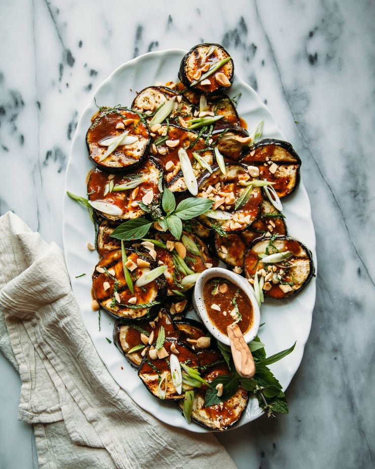 An overhead shot of grilled eggplant slices on a pale blue plate, drizzled with a deep brown sauce and garnished with chopped herbs. There is a cream coloured linen napkin nearby.