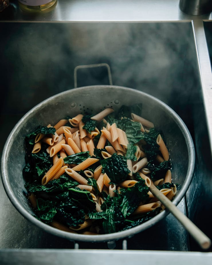 Image shows a colander full of pasta and kale draining in the sink with steam rising up from it.
