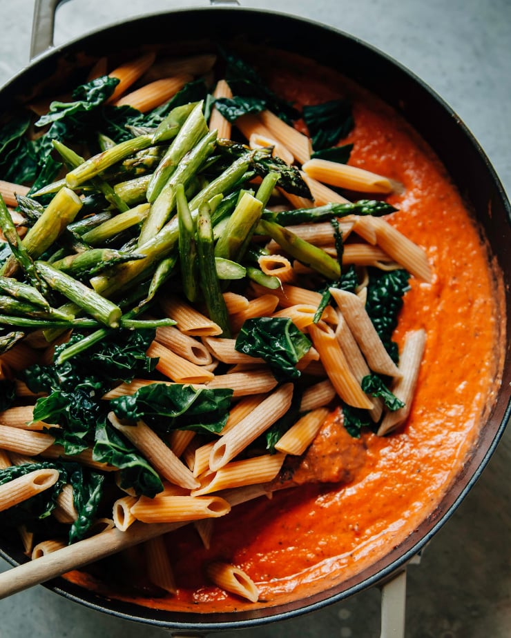 Image shows cooked pasta and vegetables being added to a red-ish pink sauce in a wide braiser-type pot.