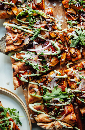 An overhead and up close shot of a flatbread covered in sauteed mushrooms, BBQ sauce, drizzles of a creamy pale green sauce, leaves of arugula, and a few chive blossoms. The flatbread is cut into squares.