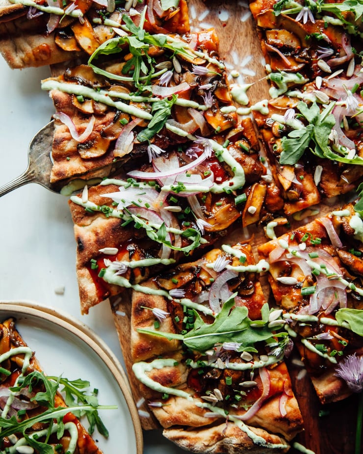 An overhead shot of a grilled flatbread with mushrooms, red onions, BBQ sauce, and a drizzle of a pale green chive sauce. The flatbread is cut into square pieces and is garnished with fresh arugula.