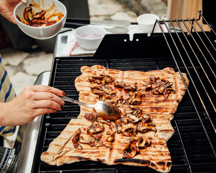 A hand is spooning saucy mushrooms onto a flatbread on an outdoor grill.