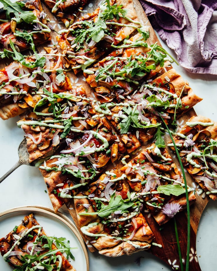 An overhead shot of a grilled flatbread with saucy mushrooms on top and a drizzle of a light green, creamy sauce. There are also leaves of arugula on top.